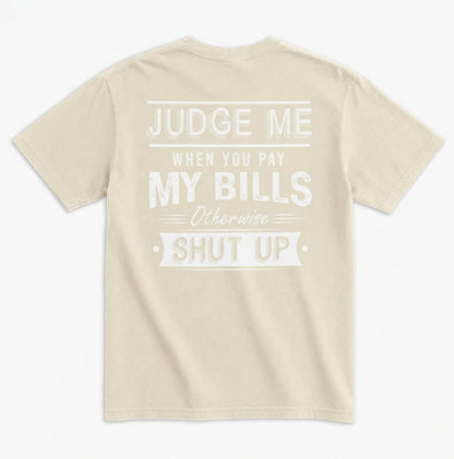 TrendyShift.Com beige unisex graphic tee with white distressed typography on the back reading Judge Me When You Pay My Bills. Otherwise SHUT UP.